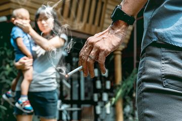 Ibu yang menggendong anaknya memarahi ayah yang memegang rokok yang berbau asap.