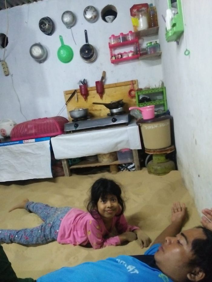 Pasir bisa memenuhi semua ruangan rumah, termasuk ruang keluarga dan dapur. Tampak seorang ayah dan putrinya tengah rebahan di kasur pasir.