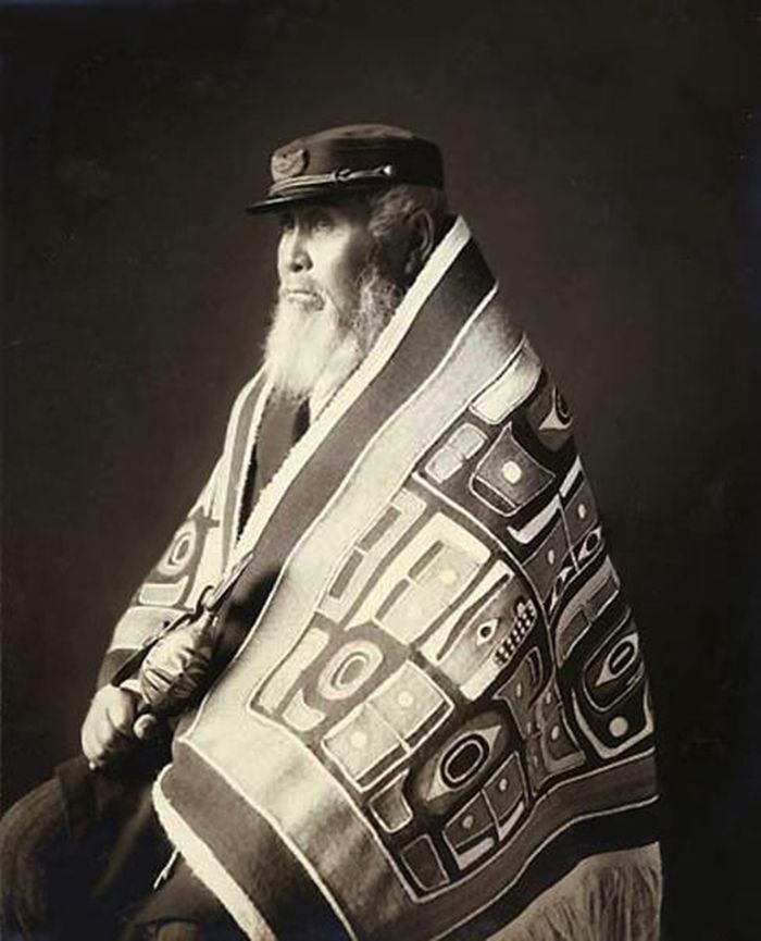 Kepala Anotklosh dari bangsa Taku. Dia mengenakan selimut tenunan Chilkan dari kulit kayu cedar dan wol kambing gunung serta topi bergaya Eropa, dan memegang mainan burung kayu berukir. Foto oleh W.H. Case, ca. 1913, Juneau, Alaska.