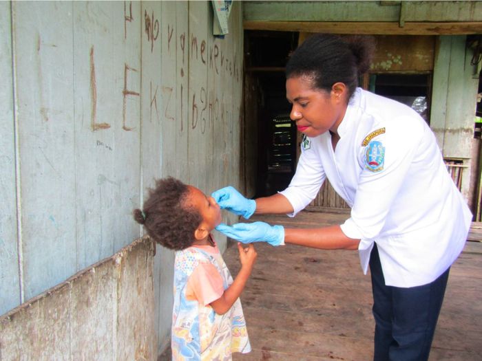 'Pemberian Vitamin Pada Anak' karya Petronela Hembring, pedagang sayuran berusia 38 tahun. Suster Magdalena Sem sedang memberikan vitamin pada seorang anak yang dikunjungi di rumahnya, ungkap Petronela dalam takarir fotonya. Sekitar 10 orang anak di Kampung Imsar mengalami ketimpangan gizi. Pemberian vitamin pada anak-anak Kampung Imsar oleh petugas kesehatan tidak dilakukan setiap bulan.