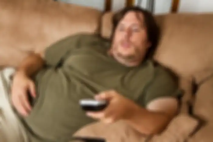 Overweight guy sitting on the couch with remote in hand trying to watch some TV.