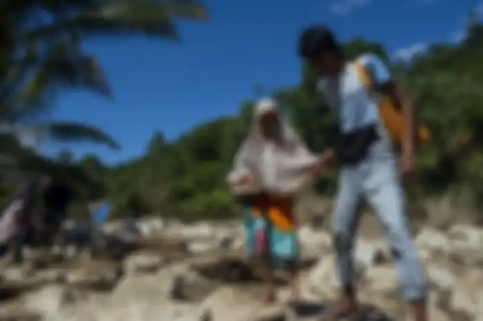 Seorang ibu dipandu saat melintasi jalan yang dipenuhi batu besar pascabanjir bandang di Desa Namo, Kecamatan Kulawi, Kabupaten Sigi, Sulawesi Tengah, Rabu (14/8/2019). Banjir bandang yang menerjang wilayah itu pada Selasa (13/8/2019) malam mengakibatkan puluhan rumah rusak berat dan hilang serta ak