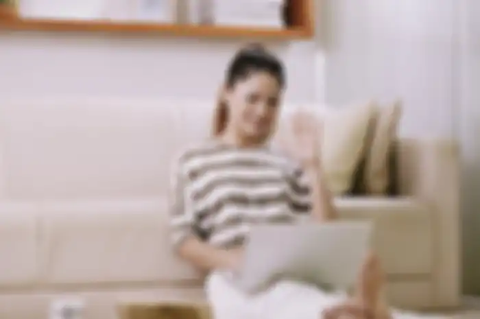 Cheerful young Asian woman sitting on the floor and video calling her friends or relatives