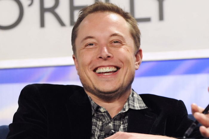Foto Elon Musk sedang tertawa lebar, Musk mengenakan baju kotak-kotak dan jas berwana hitam.