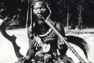 Seorang kepala perang Afrika atau seseorang yang berpangkat tinggi mengenakan pakaian upacara dengan Ngombe Ngulu di tangan kanannya.